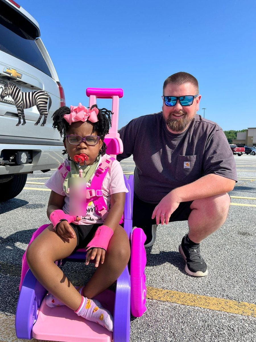 Mark kneeling beside a toddler seated in the purple and pink 3D printed mobility trainer he built